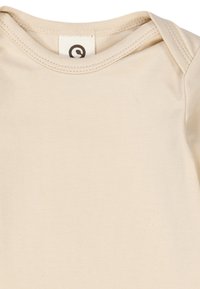 Beige puuvillainen yksiosainen haalari, jossa on pyöreä pääntie ja lyhyet hihat. Kaulassa on ympyrälogo-etiketti. Pehmeä, sileä pinta.