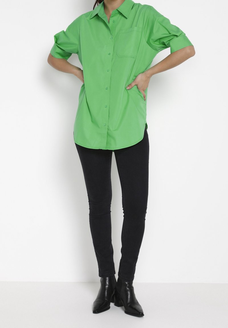 Personne portant une chemise boutonnée vert vif avec les manches retroussées, un pantalon noir skinny et des bottines noires, debout les mains sur les hanches.