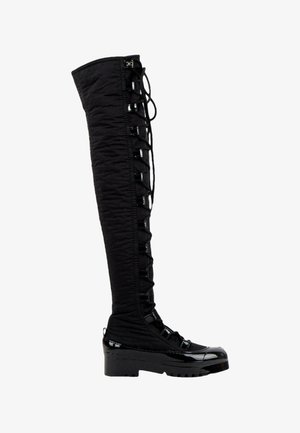 Botte noire au-dessus du genou avec devant à lacets, semelle épaisse et accents brillants sur les orteils et le talon.