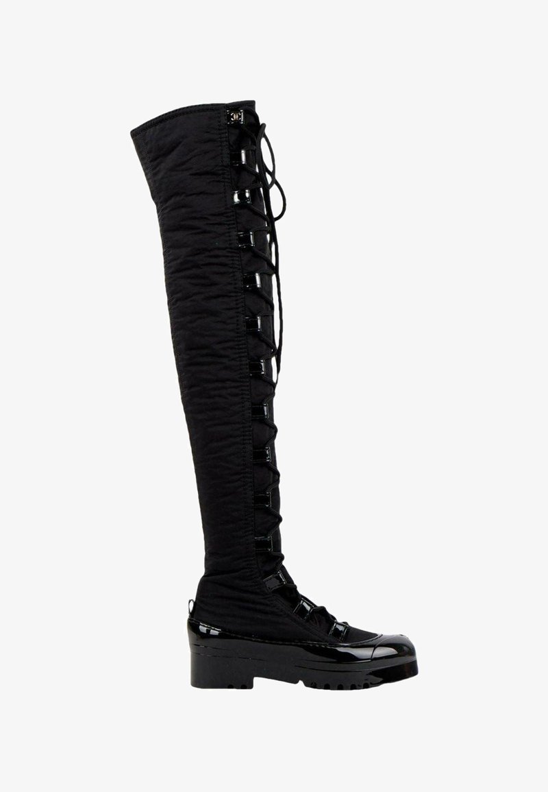 Botte noire au-dessus du genou avec devant à lacets, semelle épaisse et accents brillants sur les orteils et le talon.