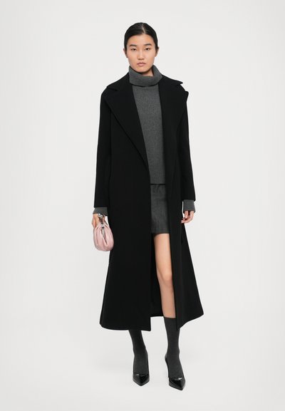 Woman wearing a long black coat over a gray turtleneck sweater and skirt, holding a small pink handbag, with gray socks and black heels.