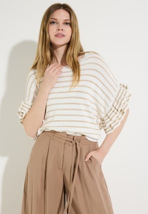 Frau mit langen blonden Haaren, die ein weiß-beiges gestreiftes Oberteil mit Rüschenärmeln und hoch taillierte beigefarbene Hose mit einem gebundenen Gürtel trägt.