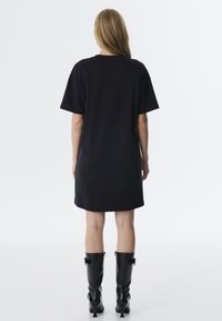 Schwarzes oversized T-Shirt Kleid mit kurzen Ärmeln, geradem Saum, weichem Stoff und lockerer Passform. Kombiniert mit knielangen, glanzenden schwarzen Stiefeln.