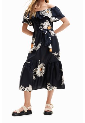 Mujer que lleva un vestido midi negro sin hombros con grandes estampados florales blancos y naranjas y sandalias crema con suelas negras.