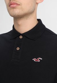 Černé polo tričko vyrobené z bavlny s límečkem a tříknoflíkovým zapínáním. Na levé straně hrudi se nachází malé vyšívané červené logo.