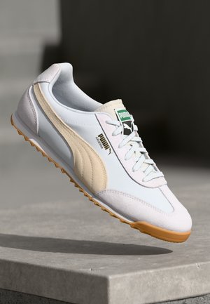 Baskets Puma blanches et beiges avec une tige en cuir lisse, une semelle en caoutchouc, un système de laçage et des détails enétiquettes vertes. Accents texturés sur les côtés.