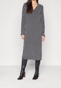 Robe en tricot avec des rayures verticales bleu marine et blanches, décolleté en V, manches longues et ourlet évasé. Associée à des bottes en cuir noires jusqu'aux genoux.