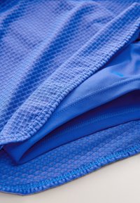 Modré vrstvy atletickej látky, vrátane sieťoviny s textúrovaným vzorom a hladkých častí, obsahujúce šijacie detaily a rôzne odtiene modrej.
