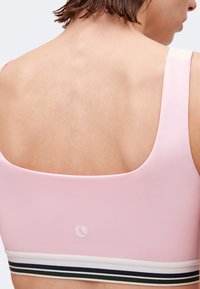 Γυναίκα με κοντά μαλλιά που φοράει ανοιχτό ροζ αθλητικό σουτιέν με μαύρη και λευκή ριγωτή ελαστική ταινία, εμφανίζεται από πίσω.