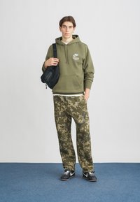 Olīvu zaļš flīsa kapuce ar priekšējo kabatu, pārī ar kamuflāžas raksta treniņbiksēm. Melna crossbody soma un tumšas kedas pabeidz izskatu.