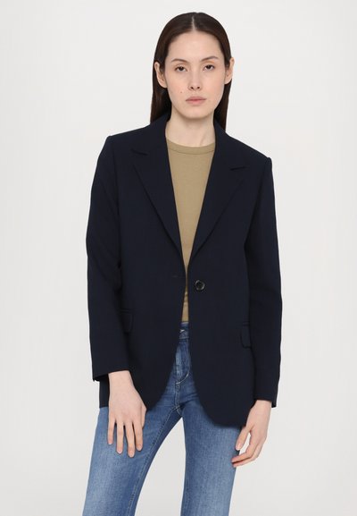 Blazer navy con cierre de un solo botón, con solapas en pico, dos bolsillos delanteros y corte relajado, llevado sobre una camisa beige y jeans.