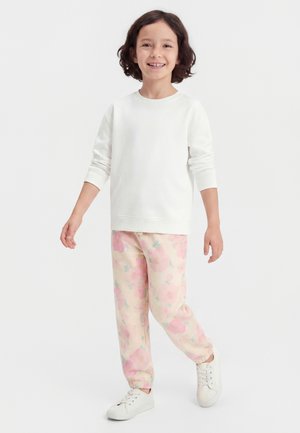 Barn med skulderlangt mørkt hår, der smiler, iført en hvid langærmet skjorte, blomstrede pink bukser og hvide sneakers, stående på en hvid baggrund.