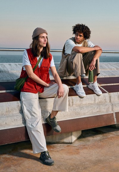 Giubbotto rosso senza maniche con tasche sopra una maglietta bianca, abbinato a pantaloni cargo beige e scarpe verdi. La seconda persona indossa un outfit beige.