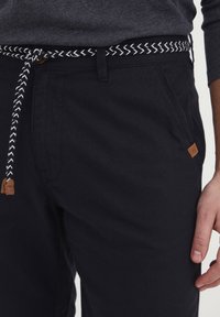 Black cotton pants with a braided white and black belt. Features a small brown leather tag on the side, smooth texture, and straight fit.