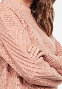 Rosa stickad tröja med en texturerad design och flätade detaljer på ärmarna samt rund halsringning. Mjukt, tjockt garnmaterial.