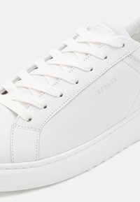 Witte leren sneaker met platte witte veters, gladde textuur, subtiele branding "CPNHGN" en een lage rubberen zool.