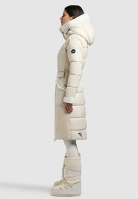 Beiger Steppmantel mit pelzgefütterter Kapuze, tailliertem Gürtel und gesteppter Textur. Kombiniert mit weißen Schneestiefeln mit Schnürung und Plateausohlen.