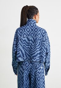 Geknipte denimjack in donker en lichtblauw met geometrische patronen, met een hoge kraag en zijzakken. Gelaagd stof met zichtbare stiksels.