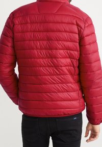 Červená prošívaná bunda s horizontálními švy, vysokým límcem a hladkou, lesklou texturou. Nenošena s černými kalhotami.