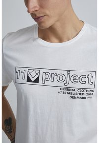 Hvit bomulls t-skjorte med et svart trykt logo som leser "11 prosjekt" og teksten "ORIGINAL CLOTHING ESTABLISHED 2020 DENMARK."