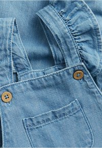 Lichtblauwe denim overall met een ruches ontwerp op de schouders, houten knopen en een voorkantzak met gestikte details.