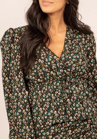 Blouse noire à motifs floraux, avec un col en V, des manches longues bouffantes et une taille ajustée ornée de fleurs colorées en blanc, jaune et vert.