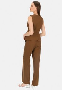 Brązowa bluzka peplum w komplecie z dopasowanymi spodniami, wykonana z gładkiego materiału. Stylizacja cechuje się dopasowanym krojem i subtelnymi detalami.