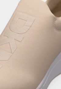 Κοντινή λήψη μπεζ αθλητικού παπουτσιού με ανάγλυφα γράμματα "DK" στο επάνω μέρος και λευκή σόλα ορατή στο κάτω δεξί μέρος.