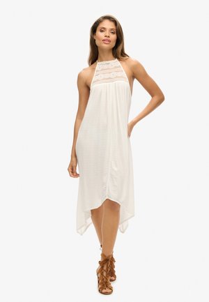 Witte halterjurk met kantdetail bij de hals, vloeiend materiaal, asymmetrische zoom en een lichte textuur, gecombineerd met lichtbruine vetersandalen.