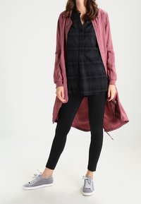 Giacca lunga rosa chiaro, blusa a quadri nera, leggins neri e sneakers grigie. Tessuto testurizzato con un design casual e dettagli con zip.