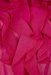 Blusa fucsia con ruches realizzata in tessuto trasparente intrecciato, con delicati dettagli e chiusura a bottone sul colletto. Texture morbida e design a strati.