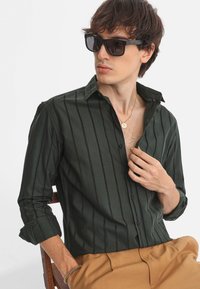Camisa de botón verde oscuro con rayas verticales negras, hecha de tela suave. Cuenta con un cuello, mangas enrollables y un corte relajado.