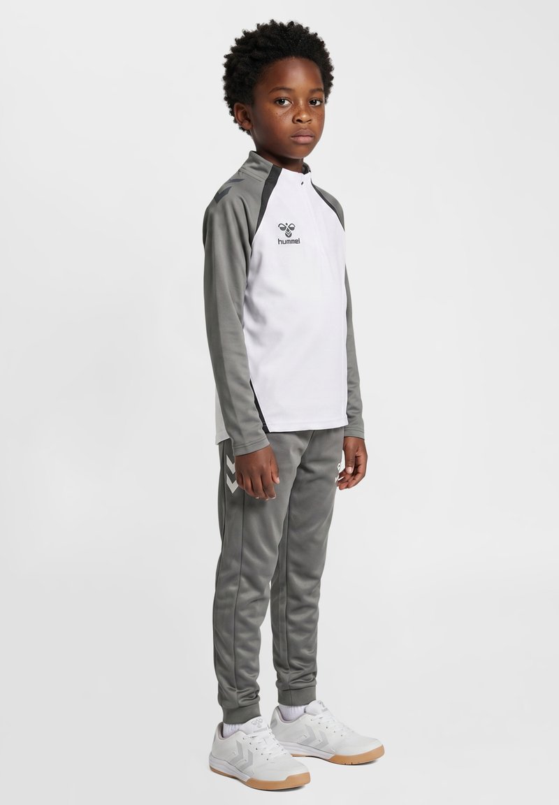 Niño con cabello rizado oscuro, vestido con una chaqueta deportiva blanca y gris de manga larga, pantalones deportivos grises a juego y zapatillas blancas, de pie de lado.