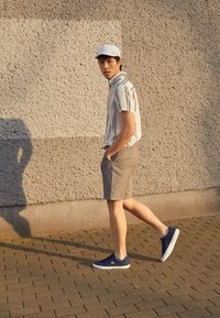 Chemise à manches courtes blanche à rayures, short gris et baskets bleu marine. Une personne debout sur un trottoir en briques avec un mur texturé en arrière-plan.