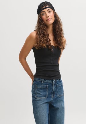 Femme aux longs cheveux bouclés portant un haut noir sans manches, un jean bleu et un bandeau noir en résille, debout les mains derrière le dos.