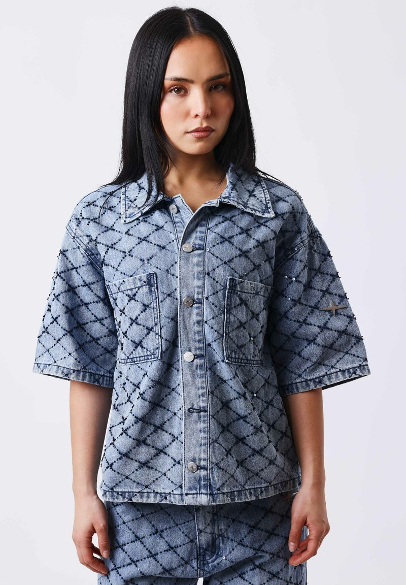 Jeune femme portant une veste en jean à manches courtes et un short assorti avec un motif en losanges foncés, debout devant un fond clair uni.