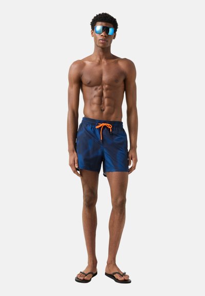 Muskularny mężczyzna w niebieskich kąpielówkach z pomarańczowym sznurkiem, czarnych klapkach i niebieskich lustrzanych okularach przeciwsłonecznych stoi na jasnym tle.