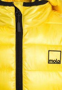 Lys gul puffjakke med sort lynlås og kraveforing, der har et "molo" logo-patch på brystet.