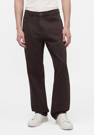 Homme portant un pantalon large marron foncé et des baskets blanches, debout sur un fond uni.