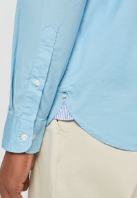 Camicia a maniche lunghe di colore azzurro chiaro con polsini abbottonati, caratterizzata da una fodera a righe all'orlo e dettagli decorativi di cucitura.