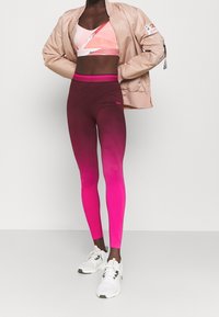 Osoba w beżowej kurtce bomber, różowo-białym staniku sportowym, gradientowych legginsach w kolorze fioletowym przechodzącym w różowy oraz białych butach sportowych, stojąca przy białej ścianie.