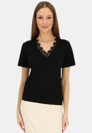 Kobieta o długich brązowych włosach, ubrana w czarną bluzkę z krótkim rękawem i dekoltem w serek ozdobioną koronką oraz beżową spódnicę, stojąca na jednolitym tle.
