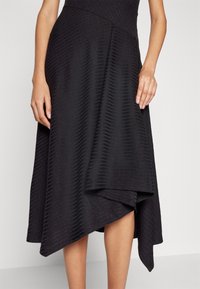 Jupe midi asymétrique noire avec des rayures horizontales texturées subtiles, portée par une personne montrant les mains et le bas des jambes.
