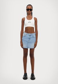 Biely skrátený top s potlačou "GCDS", svetlomodrá denimová mini sukňa a čierne masívne platformové topánky. Modelka stojí na neutrálnom pozadí.