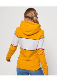 Sudadera amarilla con rayas horizontales blancas y negras, con capucha ajustable y puños acanalados. Tejido suave y corte relajado.