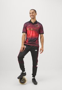 Completo da calcio rosso e nero con logo Kappa, con la scritta "TRUST IT" in lettering dorato e pantaloni patterned. Scarpe da calcio nere, in piedi su una palla.