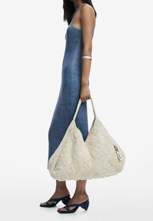 Femme portant une robe midi sans manches bleue, tenant un grand sac fourre-tout texturé de couleur beige clair, chaussée de sandales à talons bleu marine et portant un bracelet argenté au bras.