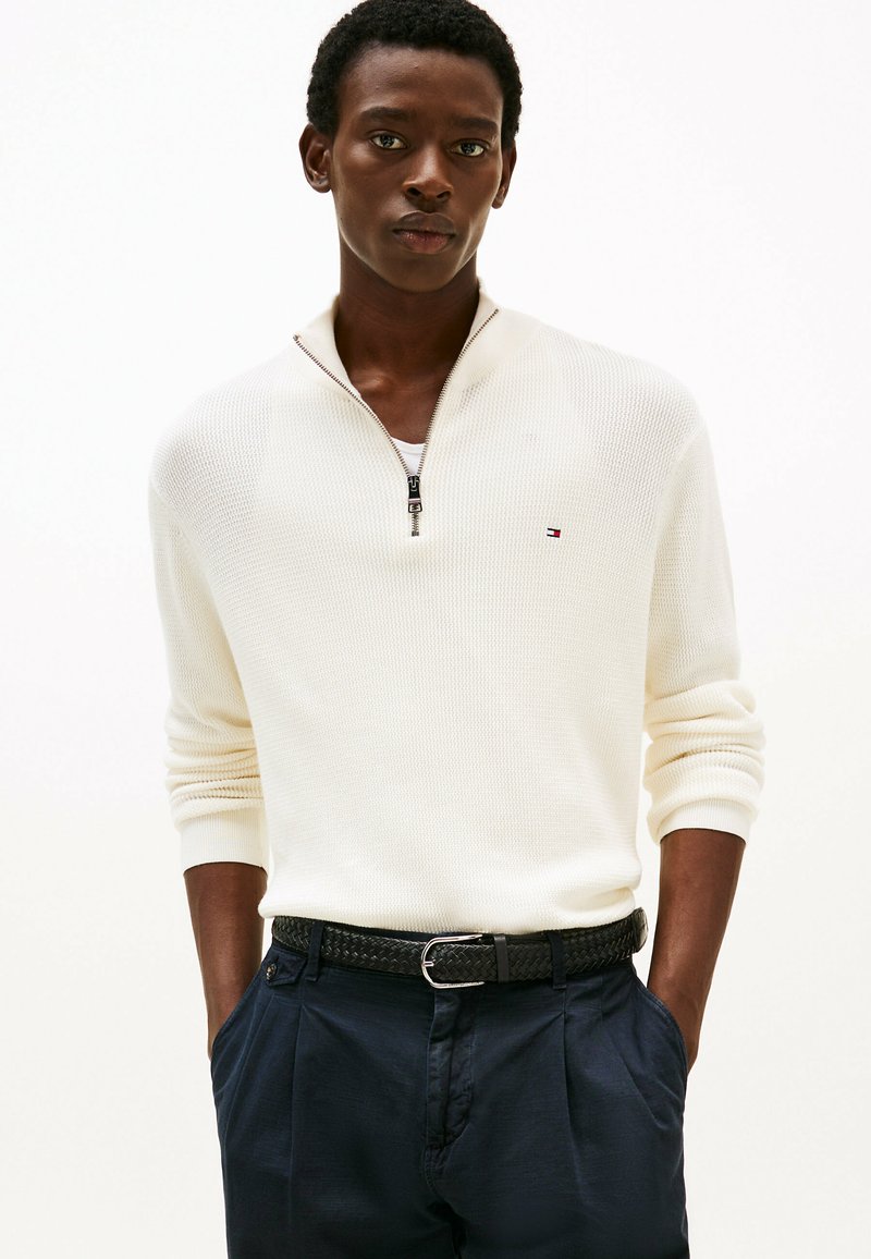 Homme portant un pull à col zippé blanc avec un petit logo, une ceinture tissée sombre et un pantalon navy à plis, les mains dans les poches sur fond blanc.