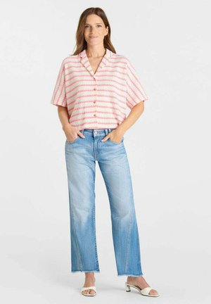 Vrouw staand met handen in zakken, gekleed in een roze-wit gestreept overhemd met knopen, lichtblauwe wijde jeans en witte sandalen met hak.