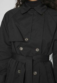 Svart trenchcoat med bältad midja, knappstängning och vikt krage. Tillverkad av mjukt, strukturerat tyg, med framfickdetalj.